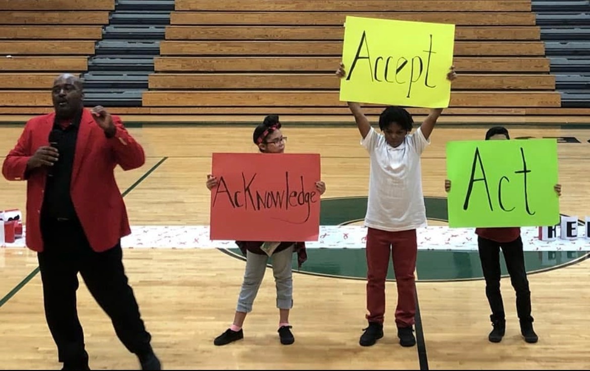On a gym court, a speaker with a microphone beside three students holding signs reading Acknowledge, Accept, and Act