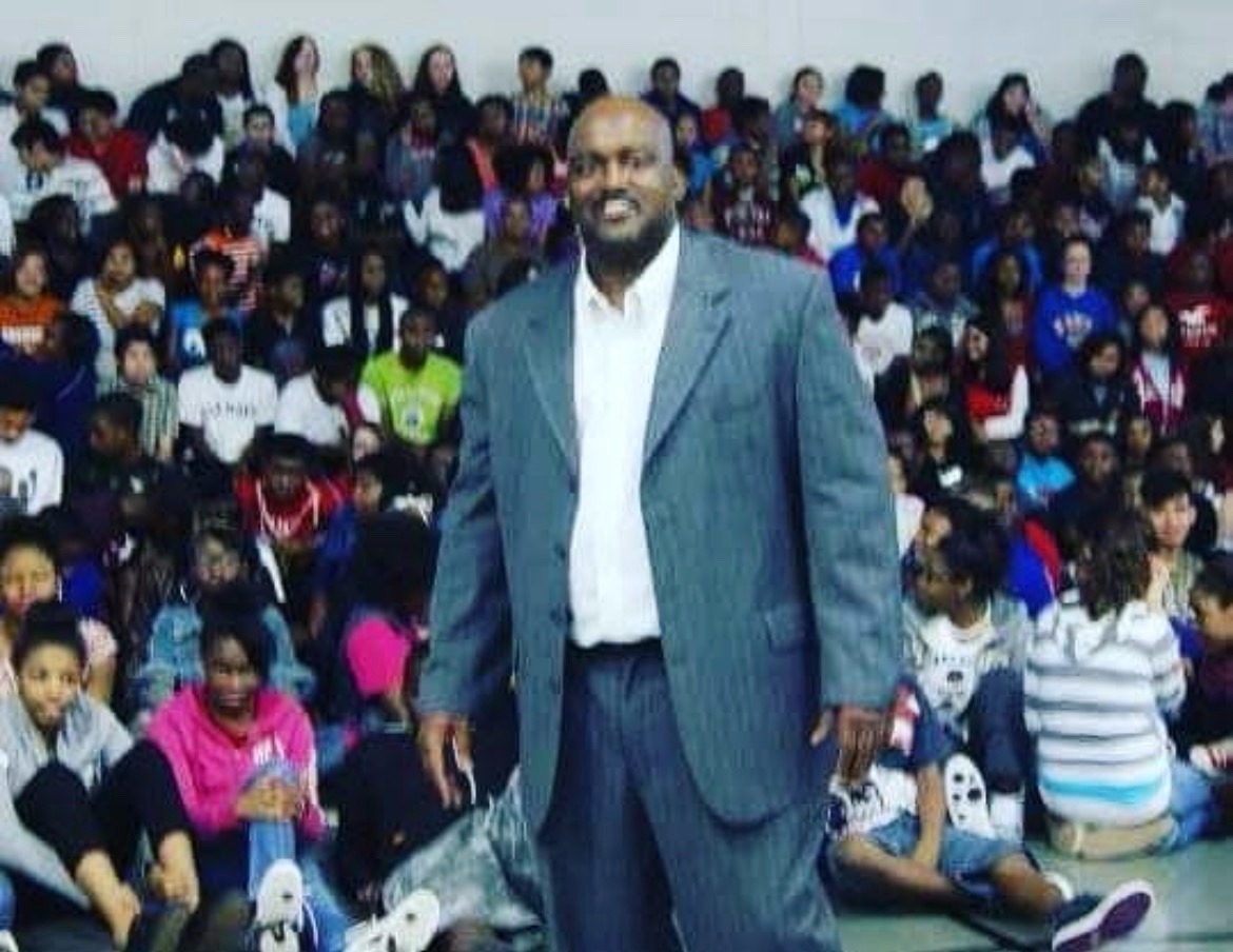 A man in a gray suit smiling in front of a large seated student assembly