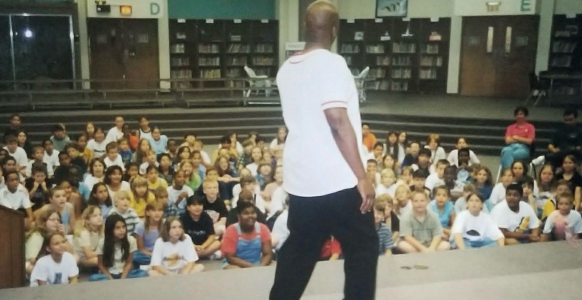 A speaker addressing a large group of students seated on the floor at a school assembly