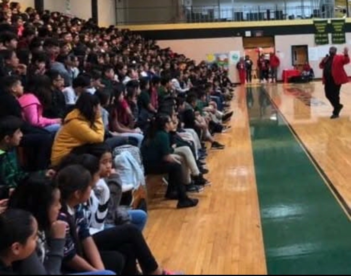 Students filling gym bleachers during an assembly, with a speaker in a red jacket on the court