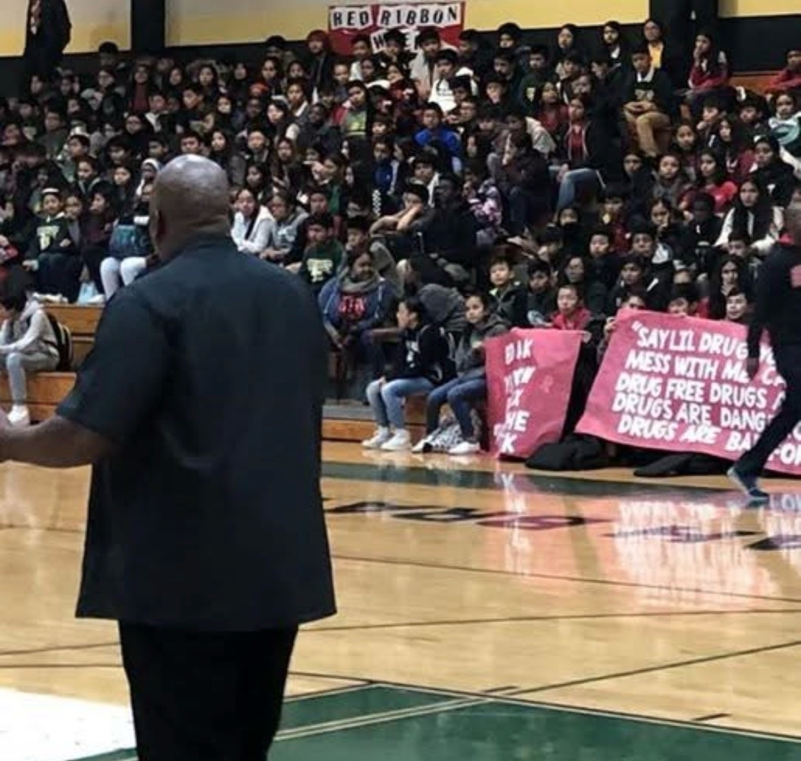 A speaker on a gym court facing students in bleachers holding drug-free banners during Red Ribbon Week